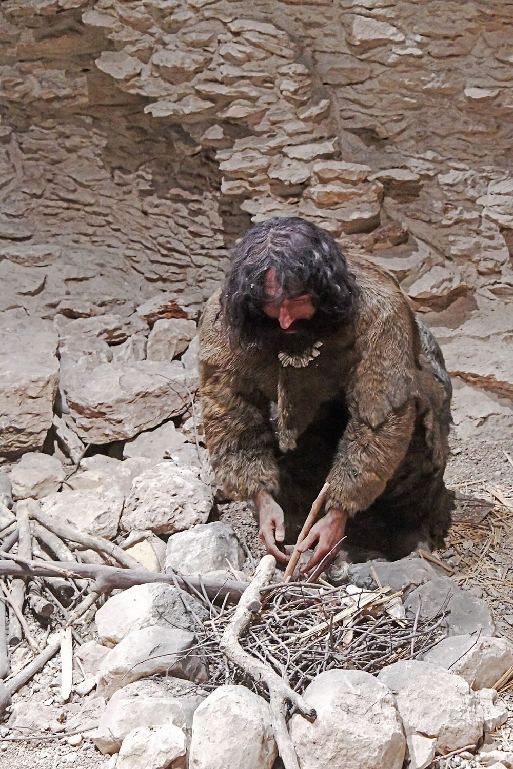 A museum reconstruction of a fur-clad hunter-gatherer kindling a fire in a rock shelter