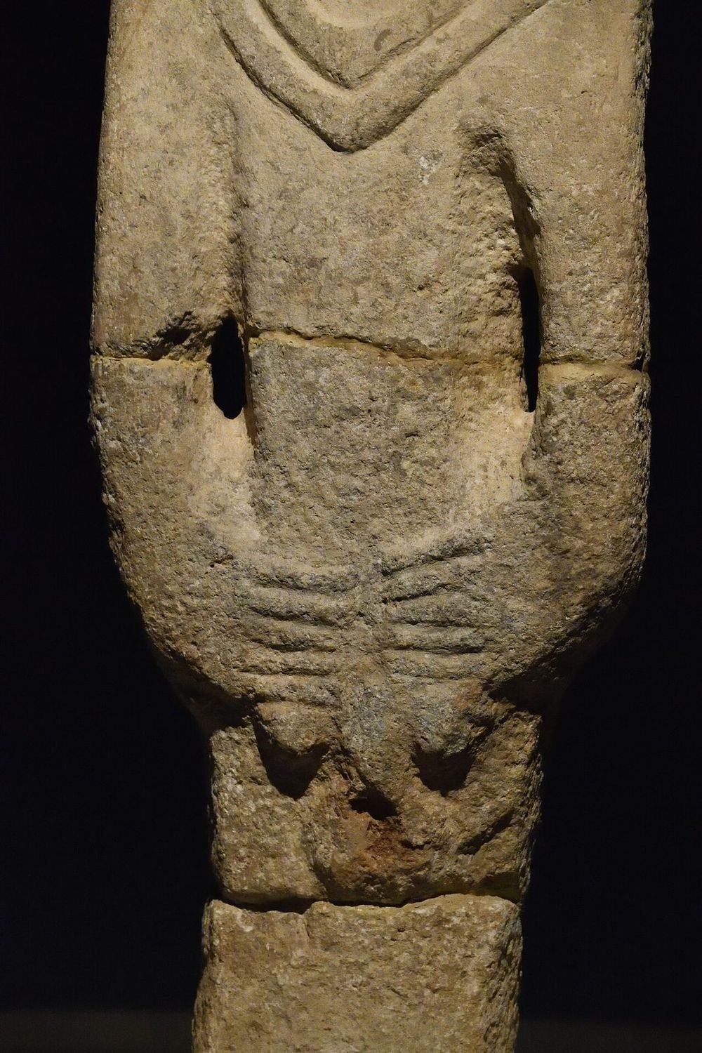 The body: a V-collar across the chest, arms carved down the sides, and both hands with fingers brought together at the front, over the genitals — the same gesture the region's T-pillars make in the abstract.